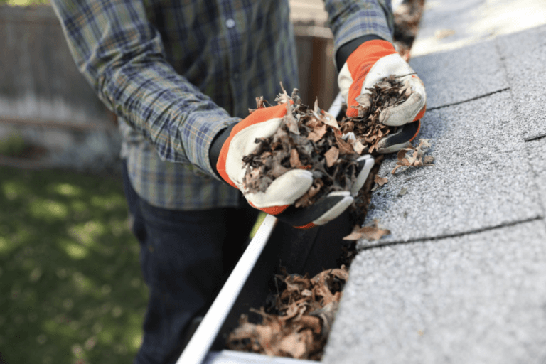 A person wearing gloves and a plaid shirt uses V-Bend Technology to remove dry leaves and debris from a house gutter along a shingled roof. The scene is outdoors with green grass and a fence in the background.